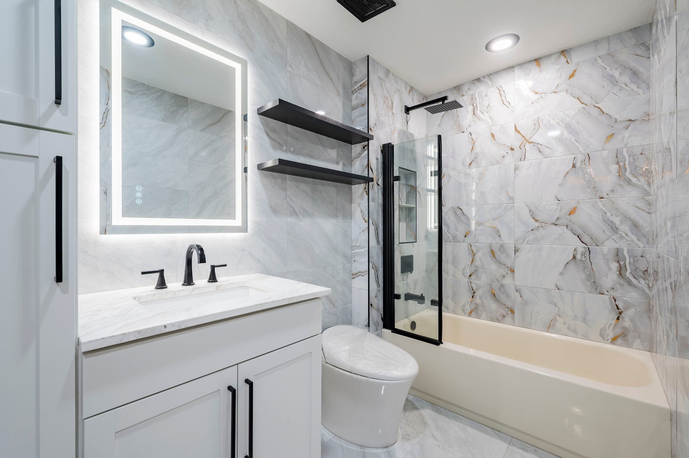 Guest bathroom at 7522 Oxford Garden Circle Apollo Beach Florida — floor-to-ceiling marble tile, LED backlit mirror, glass shower enclosure, and black fixtures