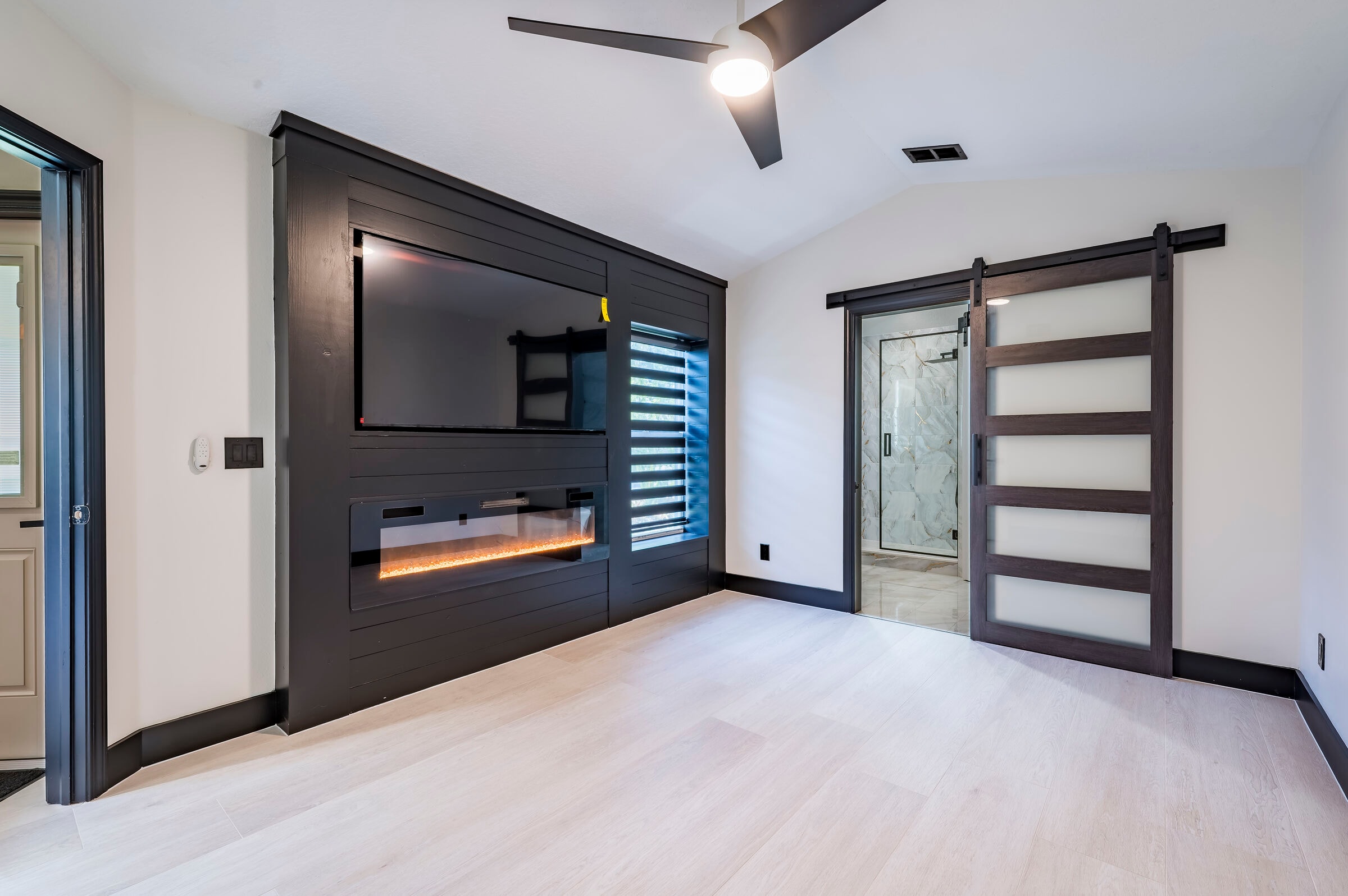 Custom primary bedroom feature wall at 7522 Oxford Garden Circle in Apollo Beach Florida — dark shiplap wall with TV mount, linear electric fireplace, and barn door to marble bath