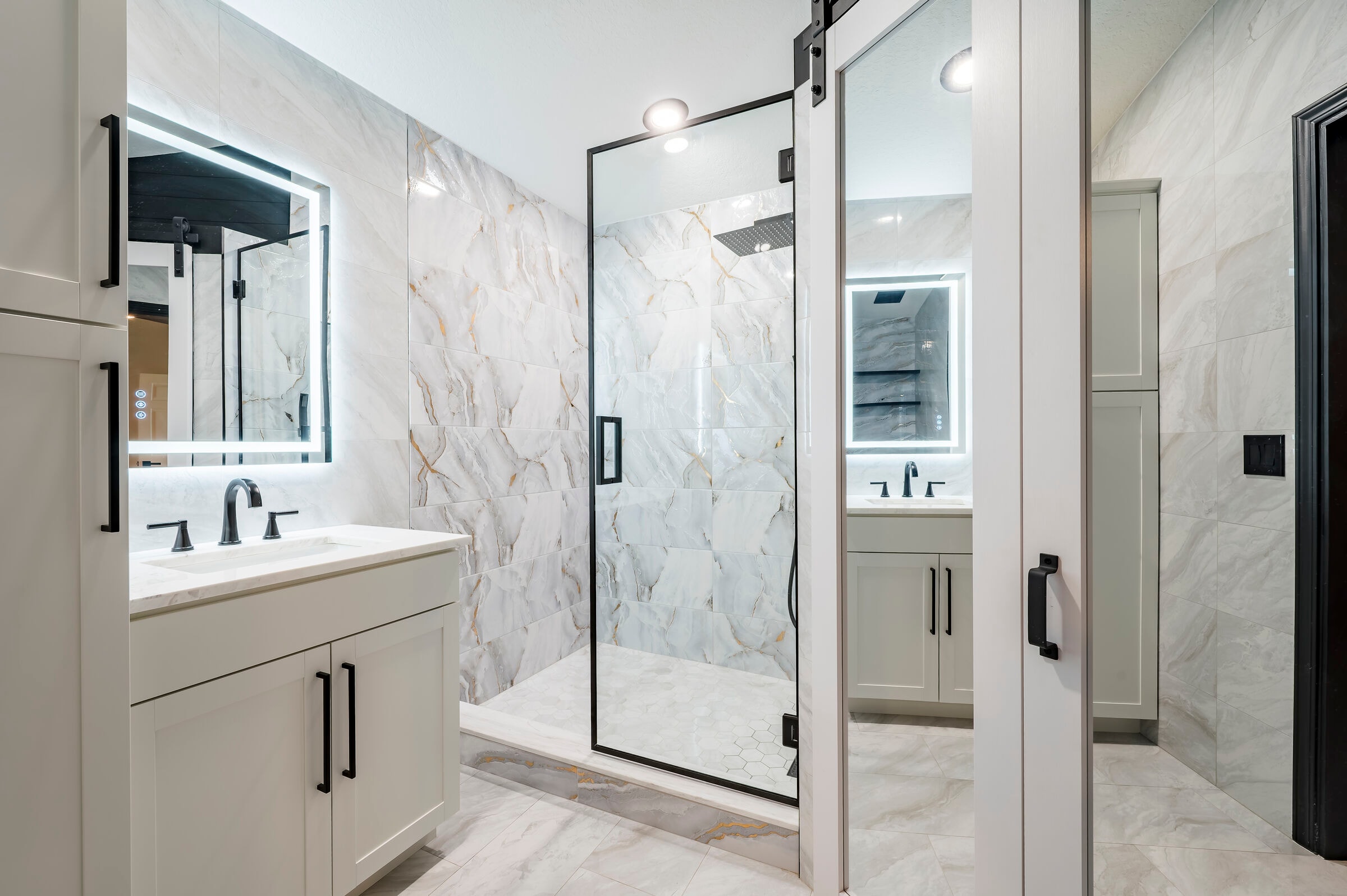 Primary bathroom at 7522 Oxford Garden Circle in Apollo Beach Florida — dual vanities, gold-veined marble tile, glass shower with barn-style door, and LED mirrors